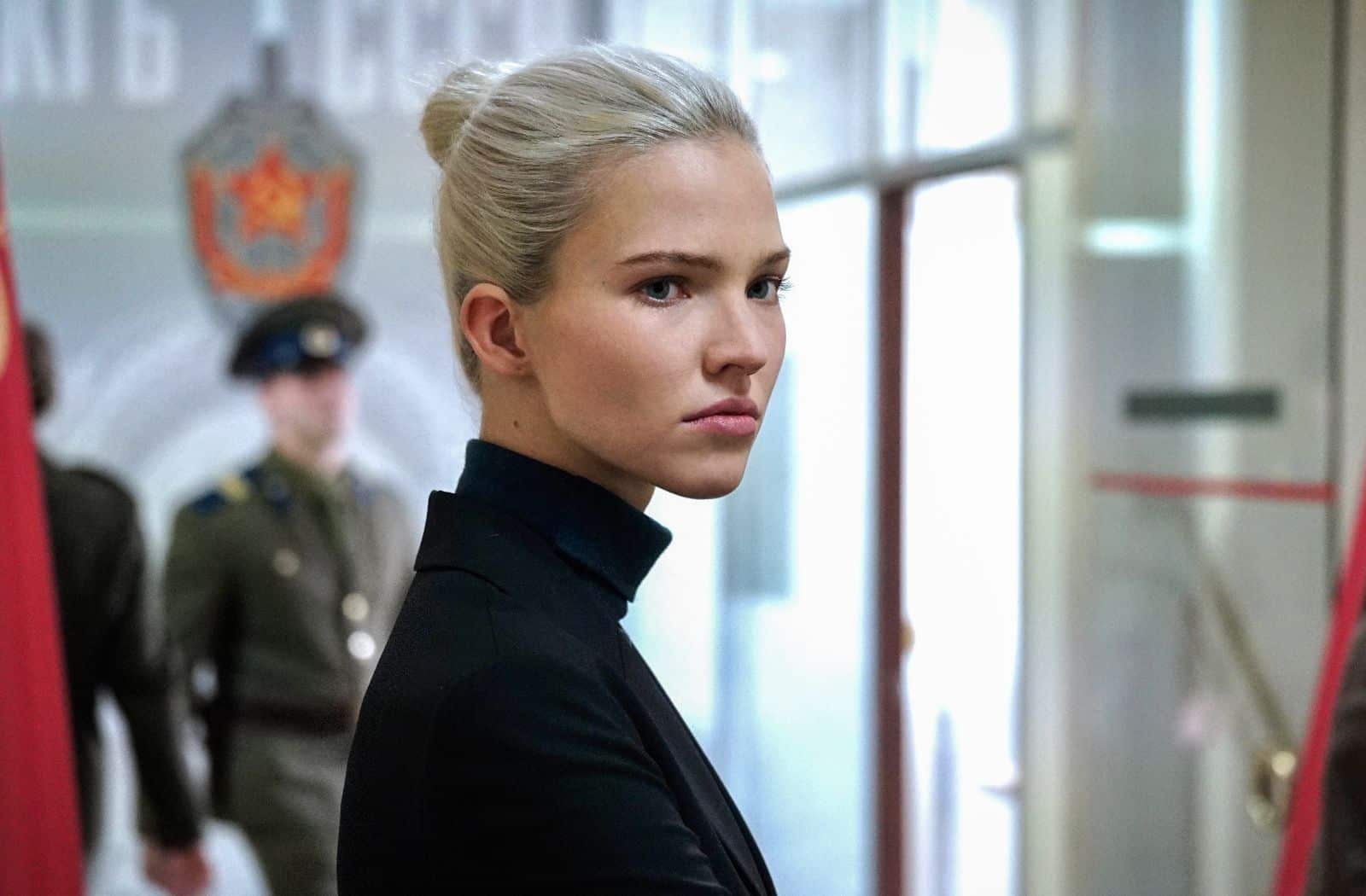 La protagonista, Anna, bajo la dirección de Luc Besson, viste ropas negras de cuello alto, lleva el pelo recogido. Mira a su derecha, mientras que a su espalda circulan dos agentes de policía rusos. 